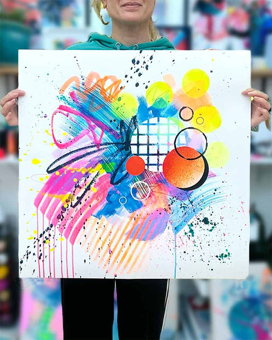 The artist Kate Elkington holding a dynamic abstract artwork featuring bold splashes of neon pink, yellow, orange, and blue, combined with graphic circle shapes, stripes, black accents and white grid patterns. Expressive brush strokes, crayon scribbles and paint drips add movement and energy, creating a vibrant and playful composition that radiates joy and positivity.
