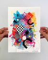 The artist Kate Elkington's hands holding a vibrant, colourful abstract artwork, featuring bold, graphic shapes, dynamic brushstrokes, and playful patterns. The composition includes splashes of neon pink, orange, bright red, and blue, layered with geometric elements such as dots, grids, and curved black lines, radiating energy and movement.