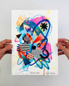 The artist Kate Elkington's hands holding a vibrant abstract artwork featuring a dynamic mix of bold shapes, patterns, and colours. The composition includes overlapping circles, triangles, and lines in neon pinks, blues, oranges, and yellows, accented with stripes, polka dots, and grid designs. The piece radiates energy and playful creativity.