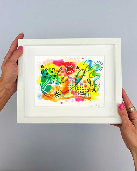 The artist Kate Elkington's hands holding a small, white, framed vibrant abstract giclée art print by Kate Elkington, featuring a playful mix of shapes and colours. Bright yellows, oranges, blues, greens, and hot pinks are blended with soft washes of watercolour, overlaid with whimsical patterns like dots, stars, and swirls. Free-flowing lines and dynamic brushstrokes create a sense of movement, while geometric elements such as squares and circles add structure. The overall composition uplifting and joyful