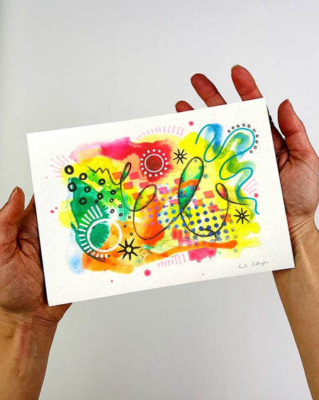 The hands of artist Kate Elkington holding an A5 sized vibrant abstract giclée art print in landscape orientation, featuring a playful mix of shapes and colours. Bright yellows, oranges, blues, greens, and hot pinks blended with soft washes of watercolour, overlaid with whimsical patterns like dots, stars, and swirls. Free-flowing lines and dynamic brushstrokes create a sense of movement, while geometric elements such as squares and circles add structure. The overall composition is uplifting and joyful.