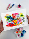 The hands of the artist Kate Elkington holding an A4 sized vibrant abstract giclée art print in landscape orientation, featuring a playful mix of shapes and colours. Bright yellows, reds, blues, greens, and pinks are blended with soft washes of watercolour, overlaid with whimsical patterns like dots, stars, and swirls. Free-flowing lines and dynamic brushstrokes create a sense of movement, while geometric elements such as squares and circles add structure. The overall composition is uplifting and joyful.
