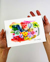 The hands of artist Kate Elkington holding an A5 sized vibrant abstract giclée art print in landscape orientation, featuring a playful mix of shapes and colours. Bright yellows, reds, blues, greens, and pinks blended with soft washes of watercolour, overlaid with whimsical patterns like dots, stars, and swirls. Free-flowing lines and dynamic brushstrokes create a sense of movement, while geometric elements such as squares and circles add structure. The overall composition is uplifting and joyful.