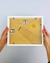 The artist Kate Elkington's hands holding a small, white picture frame, showing the back to demonstrate the fixings and the kick stand. A yellow and pink Kate Elkington logo sticker is also featured.
