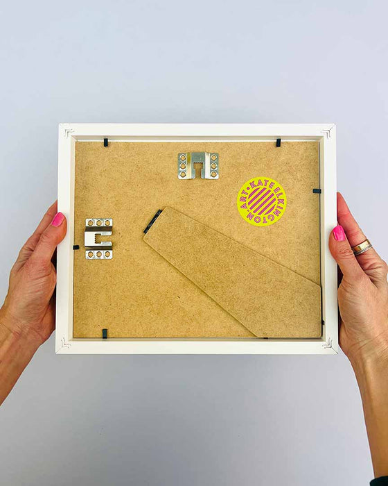 The artist Kate Elkington's hands holding a small, white picture frame, showing the back to demonstrate the fixings and the kick stand. A yellow and pink Kate Elkington logo sticker is also featured.