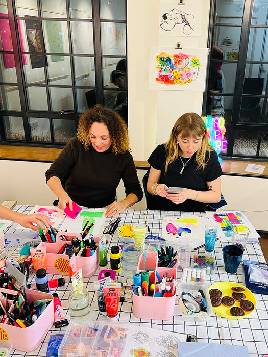 Two women sitting at a table with art supplies in a casual setting.